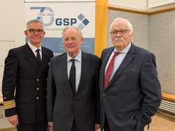 Drei ältere Männer, einer in Marineuniform, zwei in Anzügen, lächeln vor einem '70 Jahre GSP' Banner.