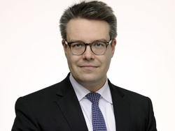 Porträt eines lächelnden Mannes mit Brille, dunklem Anzug und blau gemusterter Krawatte vor weißem Hintergrund.