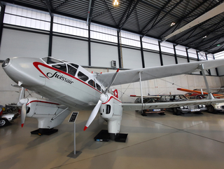Historisches silbernes Swissair-Propellerflugzeug mit Schwimmern in einem Museumshangar, dahinter Oldtimer und andere Flugzeuge.