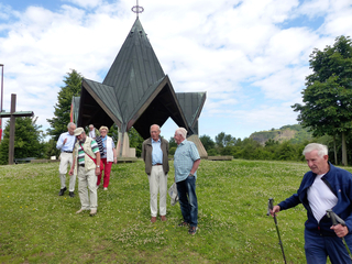 Senioren auf einer Wiese vor einer modernen Kapelle, einer mit Wanderstöcken. Bewölkter Himmel.