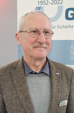 Rainer Lüttge, Sektionsleiter GSP, mit Brille, Schnurrbart und Tweed-Sakko, lächelt vor einem Jubiläumsbanner.