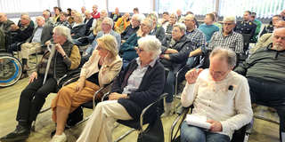 Eine vielfältige Gruppe von Erwachsenen und Senioren sitzt in einem Raum und nimmt an einer Besprechung oder Präsentation teil.