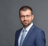 Porträt eines professionellen Mannes mit kurzen Haaren, Bart und Brille, der einen blauen Anzug und eine gestreifte Krawatte trägt.