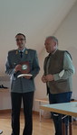 Mann in Militäruniform hält GSP-Plakette und Buch, steht neben anderem Mann in Weste.