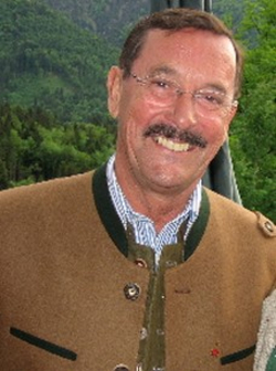 Lächelnder Mann mit Schnurrbart und Brille in einer traditionellen braunen Jacke vor grünen Bergen.