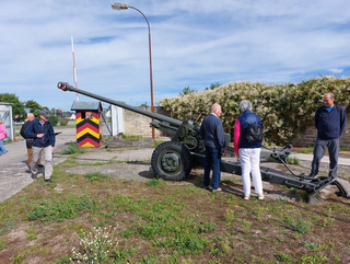 Eine Gruppe von Menschen betrachtet eine große grüne Militärkanone neben einem gestreiften Wachhäuschen und einer Schranke an einem sonnigen Tag.