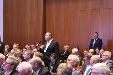 Ein Mann im Anzug spricht in ein Mikrofon vor einem Publikum in einem holzvertäfelten Konferenzraum.