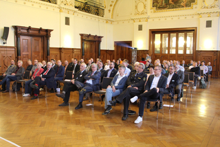 Personen bei einer Konferenz oder Präsentation in einem großen Saal mit Holzfußboden und kunstvoller Dekoration.