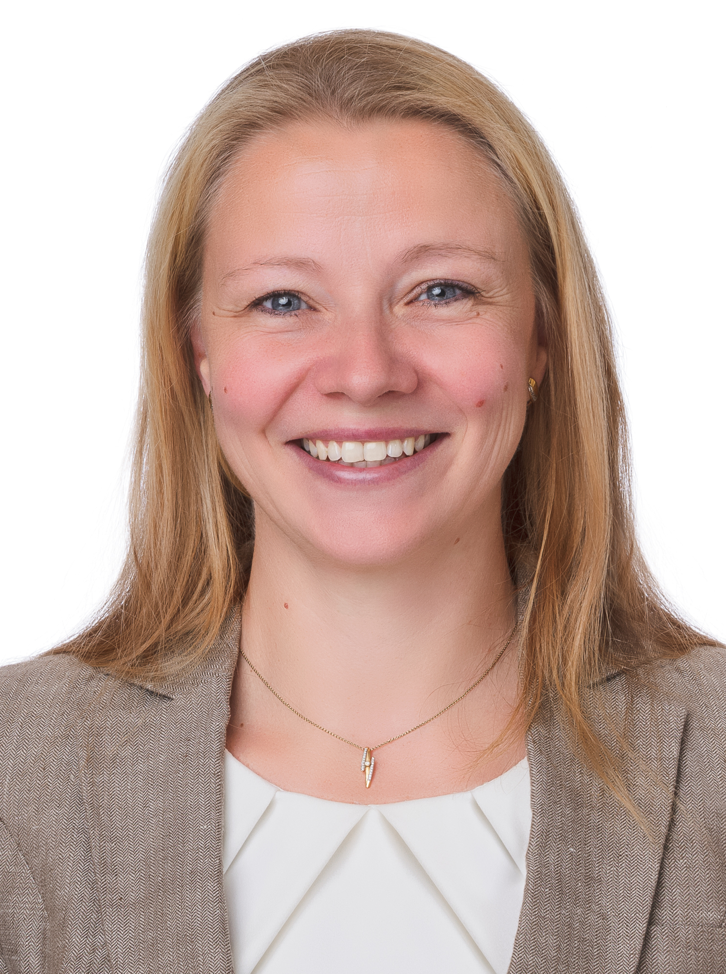 Eine lächelnde blonde Frau mit blauen Augen, einem braunen Blazer, weißem Oberteil und goldener Halskette vor weißem Hintergrund.