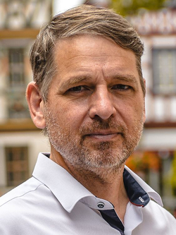 Nahaufnahme eines Mannes mittleren Alters mit kurzen braunen Haaren und Bart, der ein weißes Hemd trägt und in die Kamera blickt.