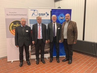 Vier Männer in Businesskleidung lächeln vor Bannern des Deutschen BundeswehrVerbandes, GSP und DWT.