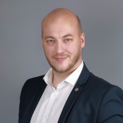 Porträt eines lächelnden, glatzköpfigen Mannes mit Bart und grünen Augen in dunklem Anzug, weißem Hemd mit Kreuzanstecker.