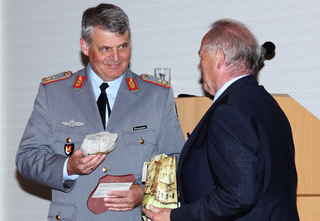 Ein lächelnder Offizier in grauer Uniform überreicht einem Mann im Anzug eine Auszeichnung (Stein und Plakette).