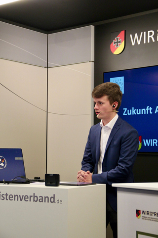 Ein junger Mann im Anzug mit Headset spricht an einem Podium bei einer Veranstaltung des Reservistenverbands der Bundeswehr.