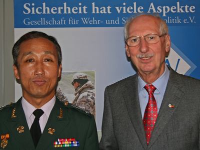Ein asiatischer Militäroffizier mit Orden und ein älterer kaukasischer Mann in Anzug und Krawatte lächeln vor einem deutschen Sicherheitsbanner.