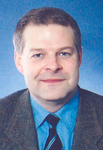 Kopfaufnahme eines lächelnden Mannes mittleren Alters mit kurzen Haaren, blauem Hemd und Krawatte vor blauem Hintergrund.
