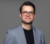 Porträt eines lächelnden Mannes mit Brille, heller grauer Jacke und dunklem Hemd vor einfarbigem grauem Hintergrund.