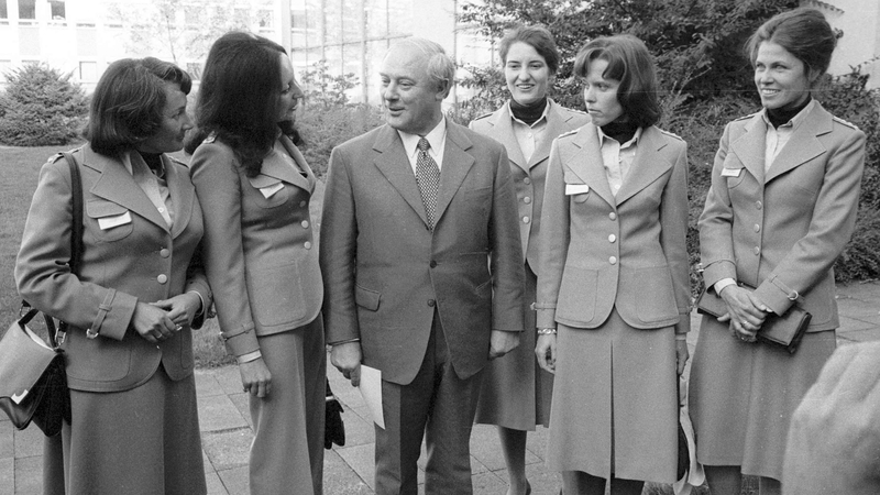 Ein Mann im Anzug unterhält sich mit fünf Frauen in 70er-Jahre-Uniformen, wahrscheinlich Flugbegleiterinnen, im Freien.