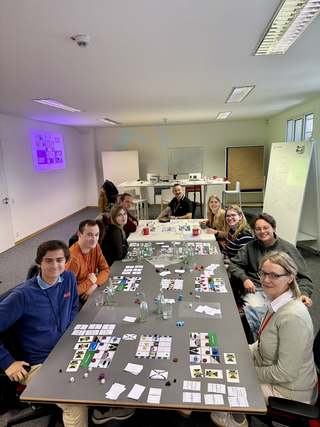 Eine Gruppe von Personen spielt ein komplexes Brettspiel mit Karten, Spielsteinen und Würfeln an einem großen Tisch im Büro.