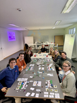 Eine Gruppe von Personen spielt ein komplexes Brettspiel mit Karten, Spielsteinen und Würfeln an einem großen Tisch im Büro.