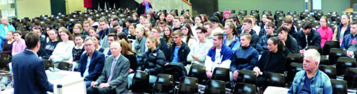 Große Gruppe von Schülern/Studenten in einem Hörsaal, die einem Vortragenden am Pult zuhören.