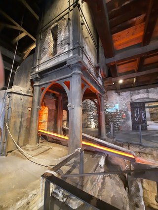 Historische Industrieanlage mit großer Ofenstruktur und hell orange-glühendem Material auf einer schrägen Rinne.