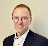 Porträt eines lächelnden, mittelalten Mannes mit kurzen Haaren und runder Brille, der einen dunklen Anzug und ein weißes Hemd trägt.