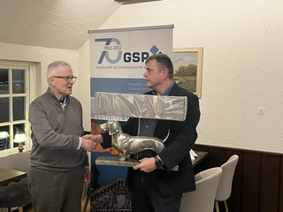 Zwei Männer schütteln Hände, einer überreicht einen Dackel-Pokal vor einem GSP-Banner zum 70-jährigen Jubiläum.