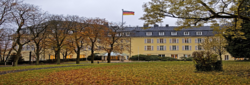 Ein großes gelbes Gebäude mit deutscher Flagge, umgeben von Herbstbäumen und herabgefallenem Laub.