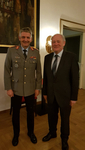 Ein lächelnder Mann in Militäruniform steht neben einem lächelnden Mann im dunklen Anzug in einem Innenraum.