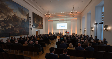 Gäste in einem großen, eleganten Saal mit historischen Wandgemälden verfolgen einen Vortrag und die "70 GSP"-Leinwand.