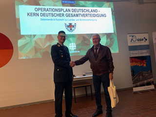 Bundeswehroffizier und Zivilist schütteln Hände vor einer Projektionswand zum Thema "Operationsplan Deutschland".