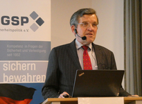 Mann mit Headset spricht an Rednerpult vor GSP-Banner über Sicherheits- und Verteidigungspolitik, Laptop auf Pult.