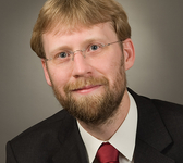 Porträt eines lächelnden Mannes mit blonden Haaren, Bart, Brille, Anzug und roter Krawatte vor grauem Hintergrund.