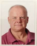 Kopfaufnahme eines kahlen, älteren Mannes mit hellem Teint in einem rosa Poloshirt, der direkt in die Kamera blickt.