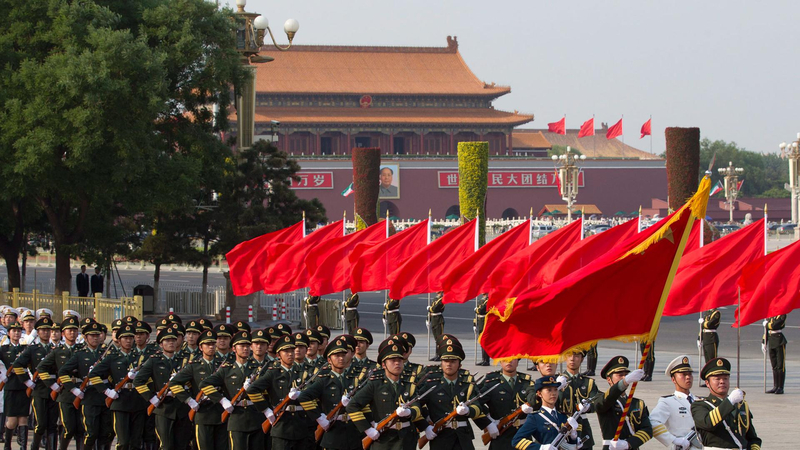 Chinesische Militärparade: Uniformierte Soldaten marschieren mit Gewehren und roten Flaggen vor dem Tiananmen-Tor mit Mao-Porträt.