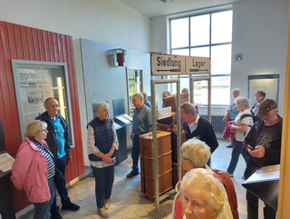 Ältere Besucher in einer Ausstellung, die einen Wegweiser 'Siedlung | Lager' und einen Stapel alter Koffer zeigt.