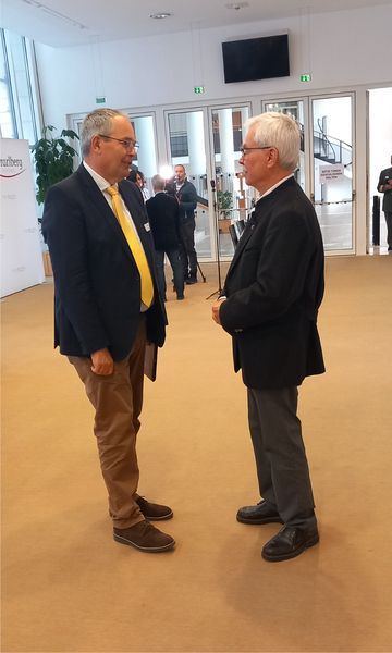 Zwei Männer, einer mit gelber Krawatte, einer in dunkler Jacke, unterhalten sich in einer Konferenzhalle mit Kamerateam im Hintergrund.