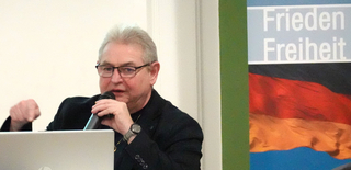 Älterer Mann mit Brille spricht in Mikrofon vor Banner mit "Frieden Freiheit" und deutscher Flagge.
