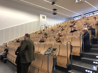 Blick in einen modernen Hörsaal mit großem Publikum, das in Reihen von hellen Holzstühlen sitzt, und hoher Decke.