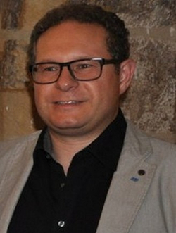 Lächelnder Mann mit Brille, dunklen Locken, schwarzem Hemd und hellem Sakko.