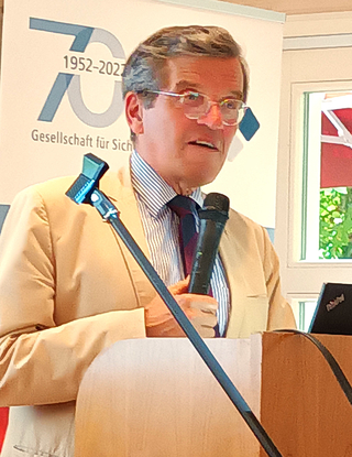 Mann spricht an einem Rednerpult mit Mikrofon. Banner im Hintergrund zeigt '70 Jahre 1952-2022'.