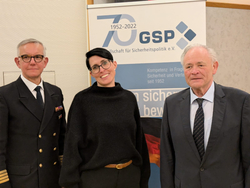 Drei Personen, zwei Männer und eine lächelnde Frau, stehen vor einem GSP-Banner zum 70-jährigen Jubiläum der Gesellschaft für Sicherheitspolitik.