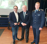 Drei Männer: ein Sprecher mit Mikrofon, ein Herr im Anzug und ein Offizier in Uniform, bei einer Veranstaltung mit 'FEUERWEHR'.