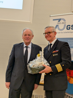 Zwei lächelnde Männer, einer im Anzug, der andere in Marineuniform mit Medaillen, bei 70-Jahr-Feier mit Geschenkkorb.