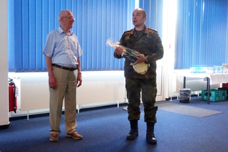 Zwei Männer, ein Zivilist und ein Militär in Uniform mit Geschenk, der spricht, drinnen vor blauen Jalousien.