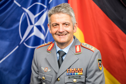 Lächelnder deutscher Militäroffizier Sollfrank in Uniform mit Orden, vor NATO- und Deutschlandflaggen.