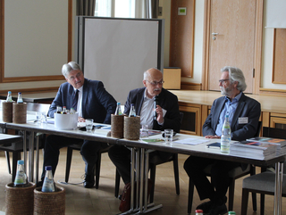 Drei Männer an einem Konferenztisch, einer spricht in ein Mikrofon, Getränke und Papiere auf dem Tisch.