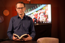 Ein Mann mit Brille und Dutt hält ein offenes Buch. Im Hintergrund zeigt ein Fernseher jubelnde Sportfans.