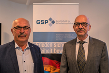 Zwei Männer mit Brillen und Schnurrbärten in Anzügen vor GSP-Banner und Deutschlandflagge.